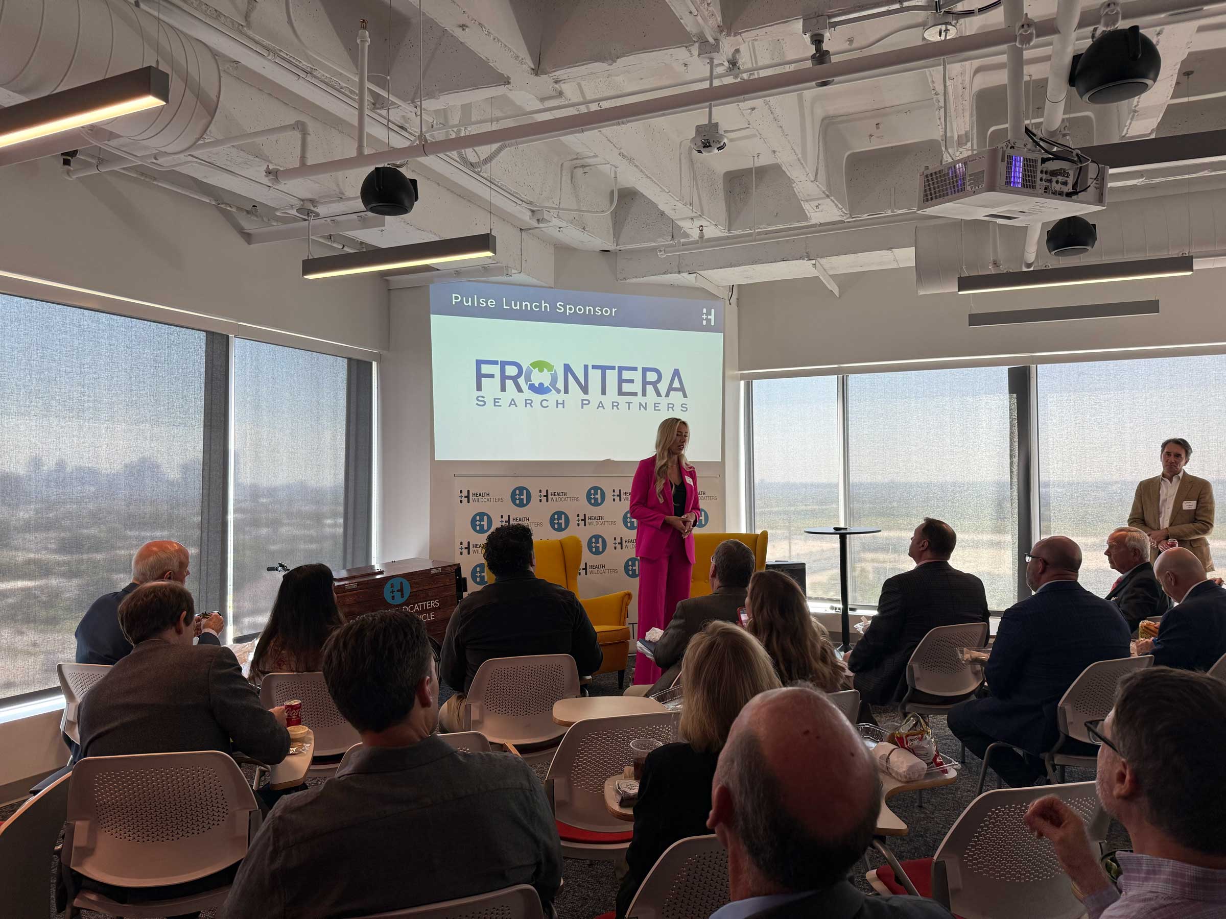 Woman presenting at a professional healthcare event with a screen displaying “Pulse Lunch Sponsor Frontera Search Partners,” highlighting industry thought leadership and partnerships.