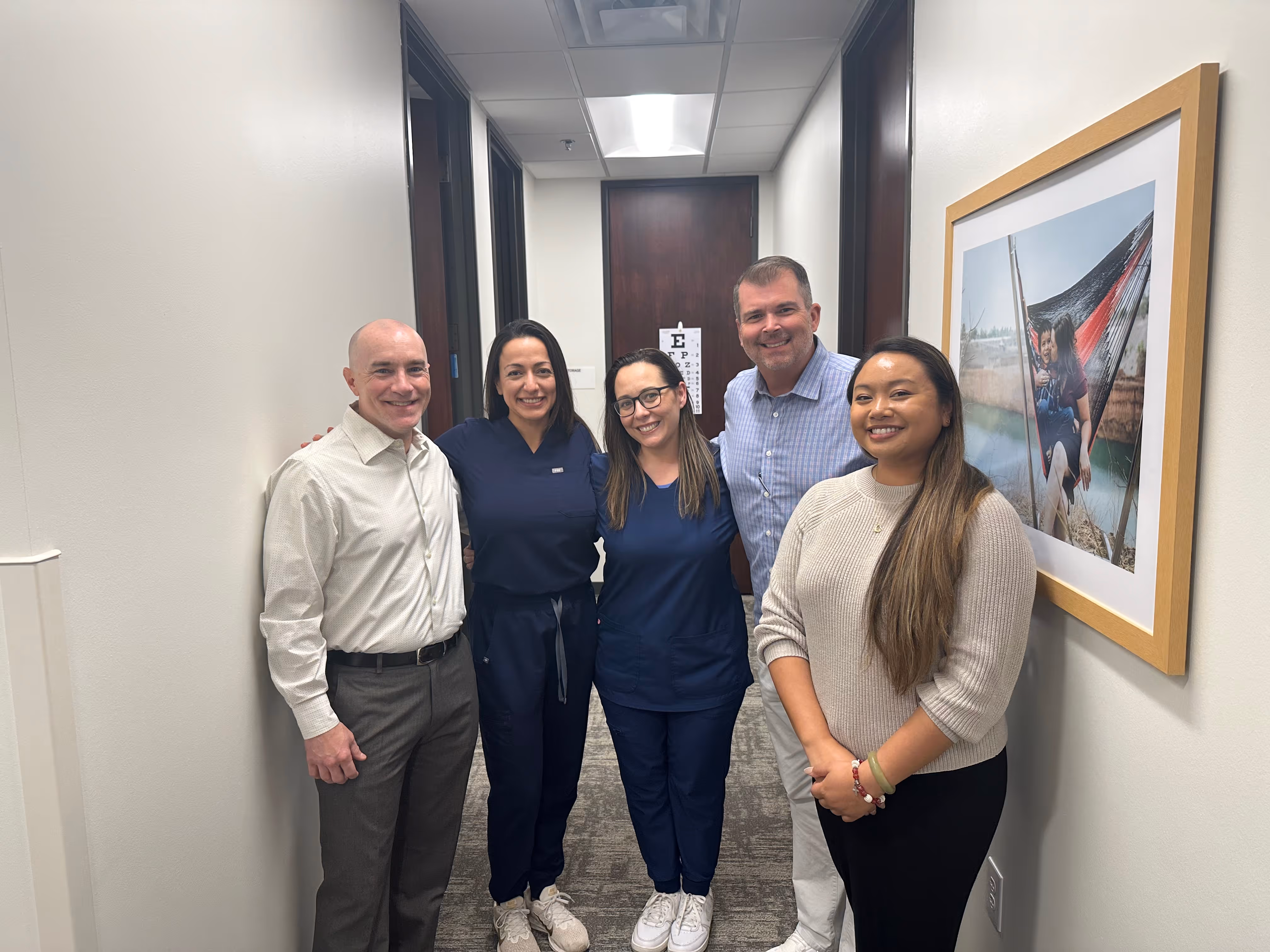 Group of healthcare professionals and staff standing together in a hallway, including clinicians in scrubs and team members in business attire, representing interdisciplinary collaboration.