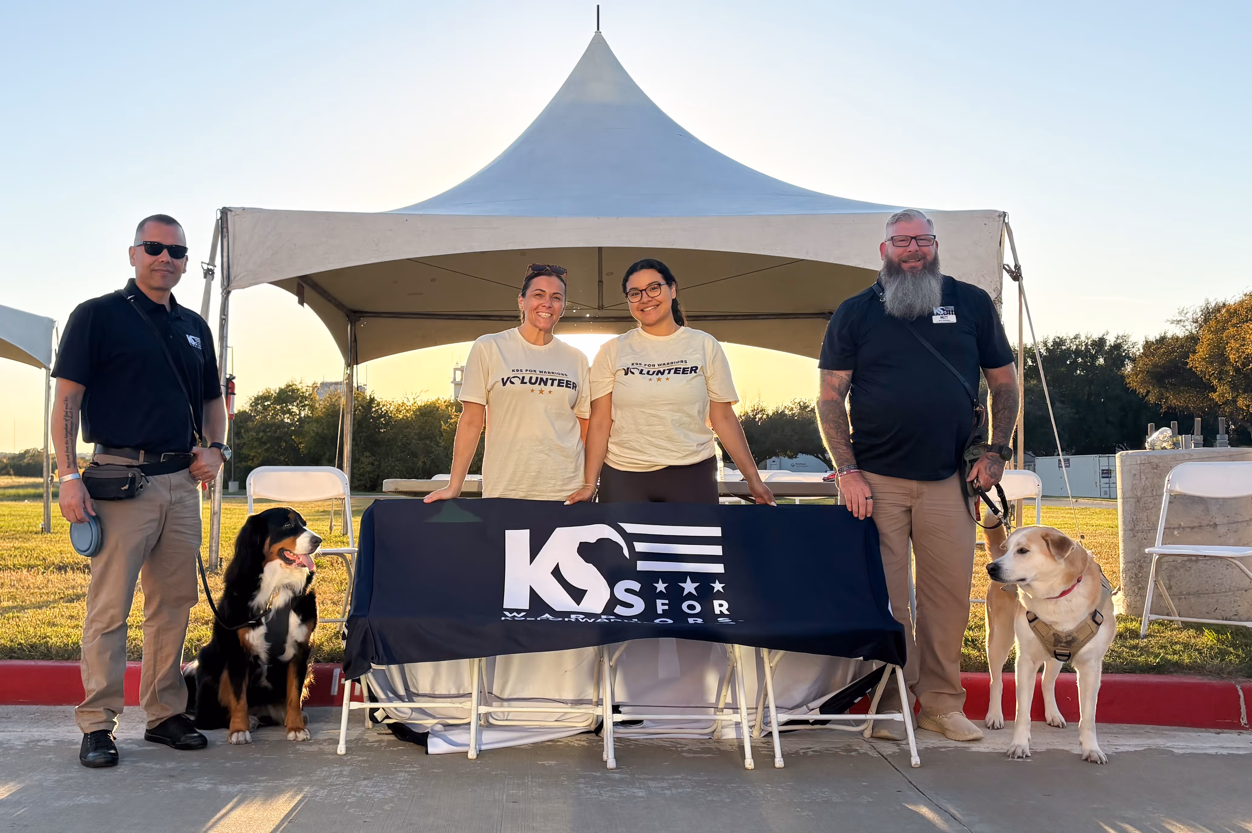 Volunteers standing with service dogs in front of a tent, holding a K9s For Warriors banner, highlighting veteran support and nonprofit community involvement.