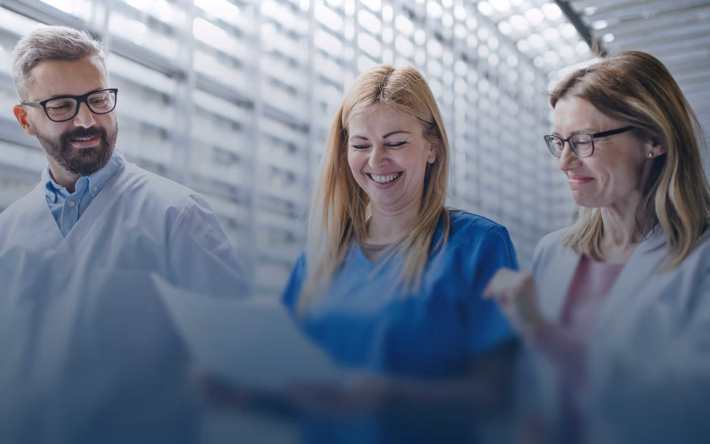Three healthcare professionals in lab coats and scrubs reviewing documents together in a modern medical facility, representing coordinated patient care and clinical collaboration.