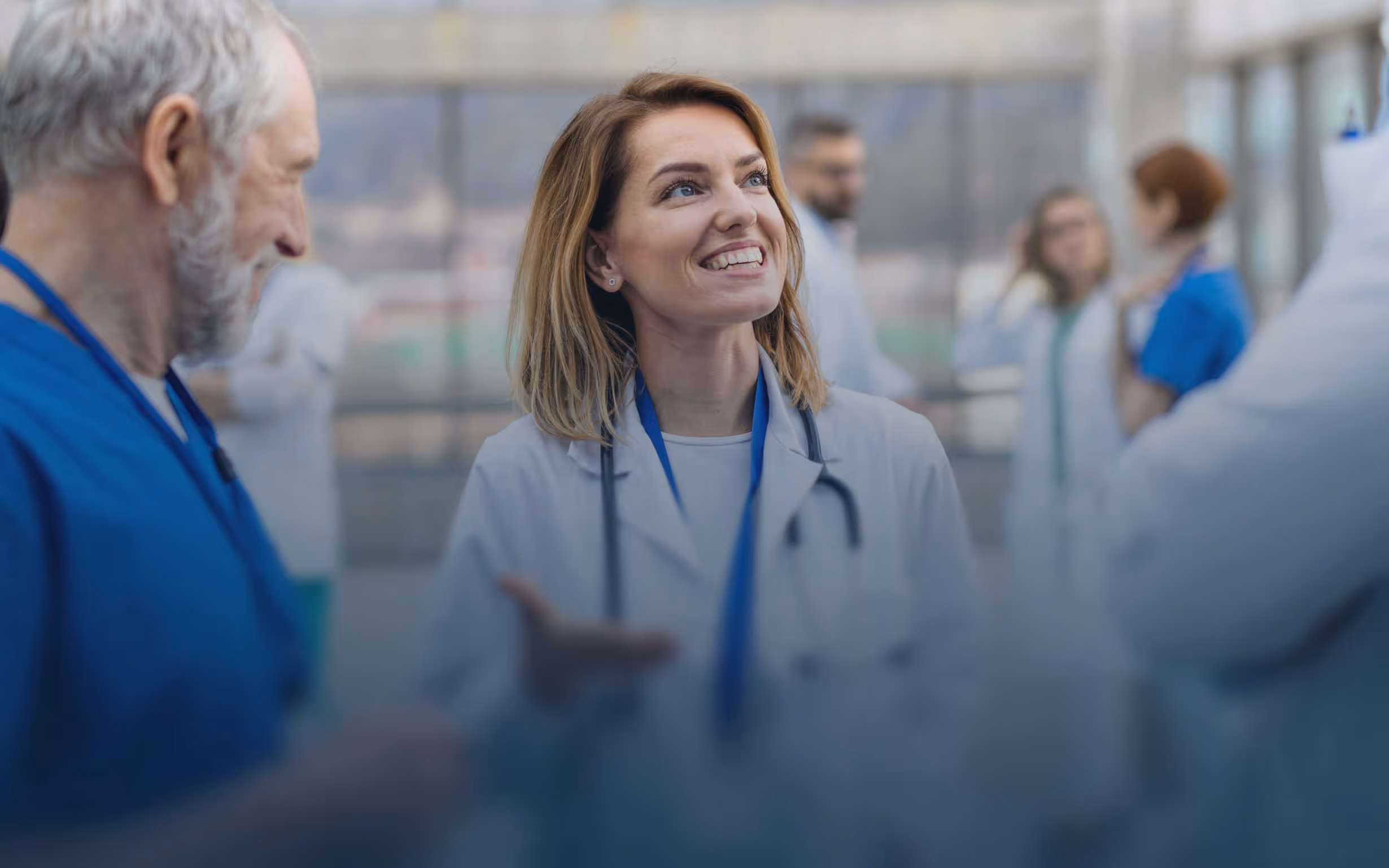 Smiling female doctor wearing a stethoscope speaking with colleagues in a hospital corridor, illustrating teamwork and collaboration among healthcare professionals.