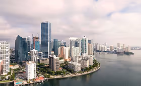 Aerial view of a coastal city skyline featuring modern high-rise buildings along a curved waterfront under a cloudy sky.