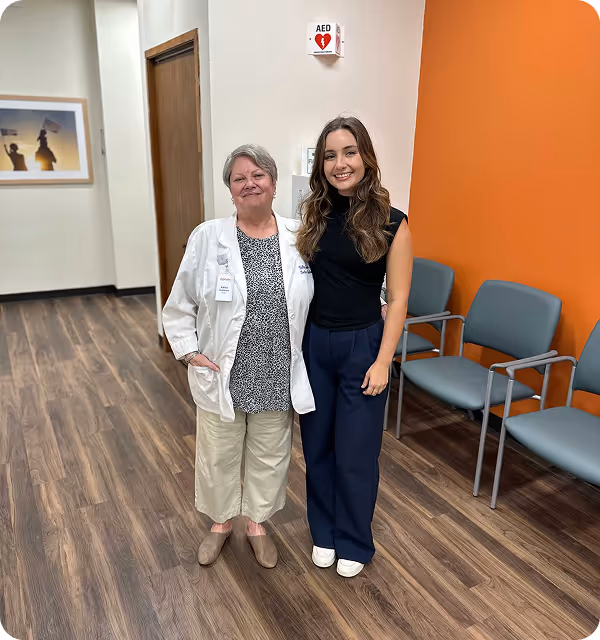 Two women standing in a modern waiting area, one in a white medical coat and the other in business attire, representing patient care and administrative collaboration.