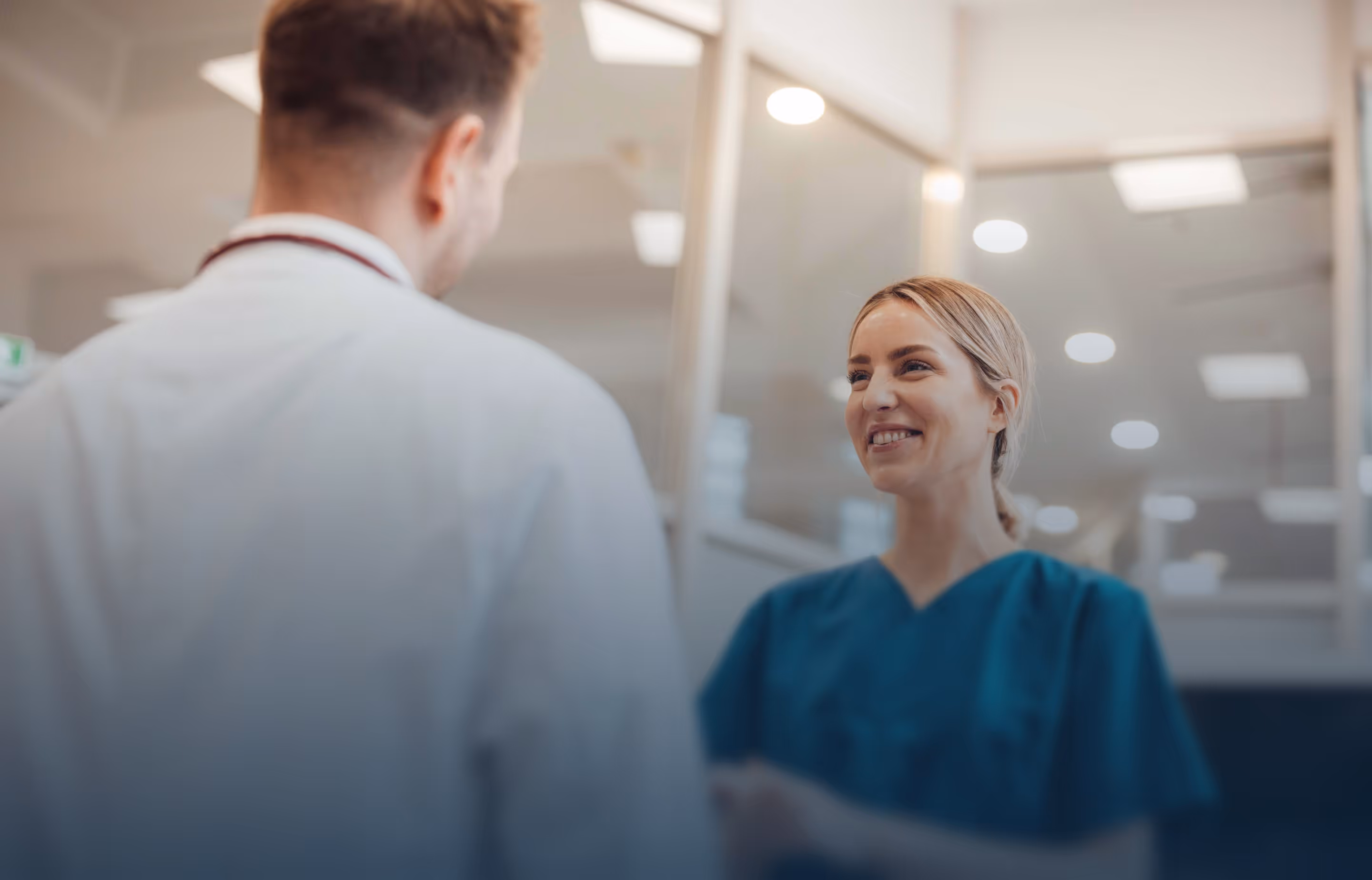Female nurse in teal scrubs speaking with a male doctor wearing a stethoscope in a hospital setting, illustrating collaboration between nursing and medical staff.