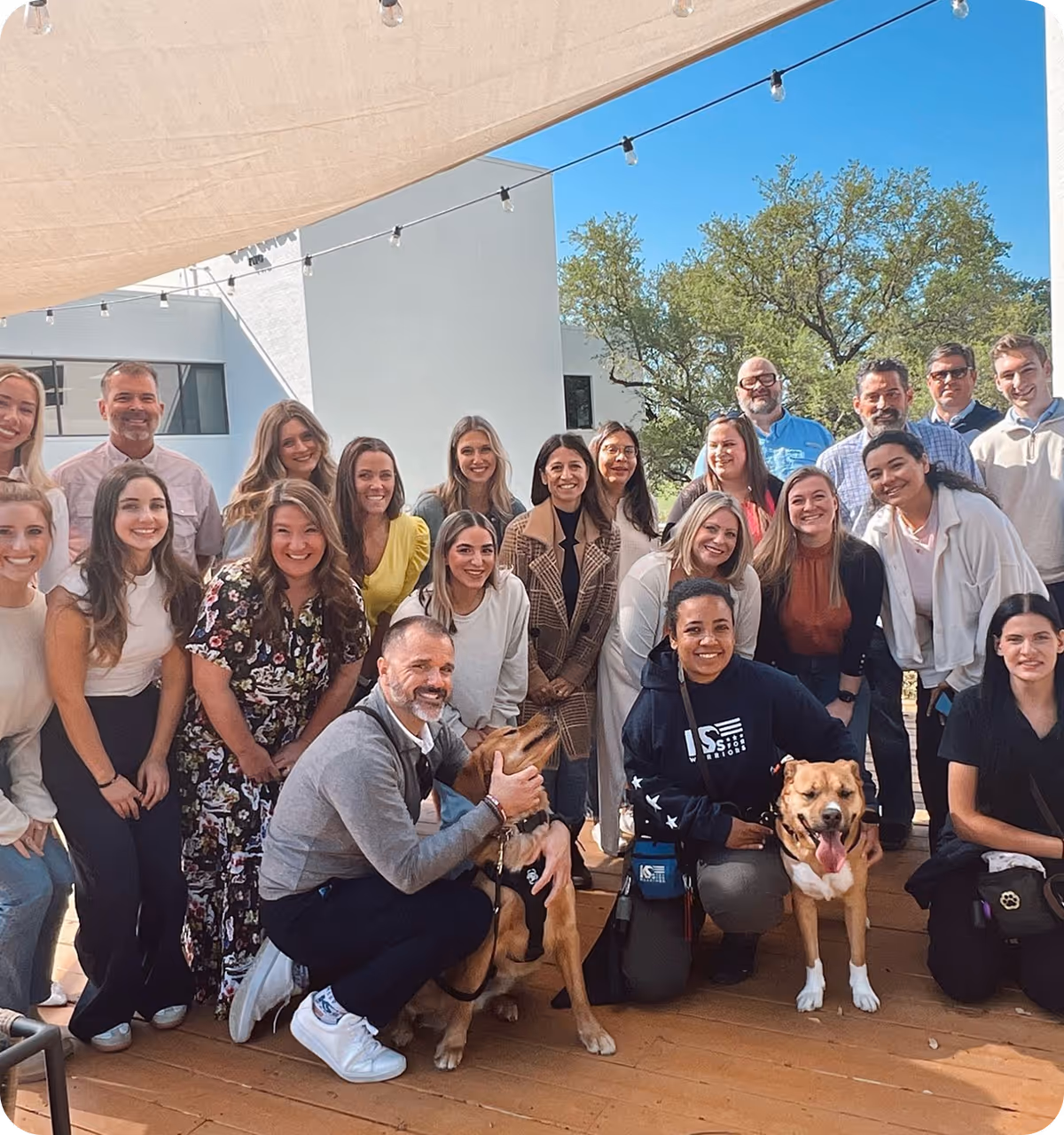 Group of smiling people posing outdoors with two dogs on a wooden deck under string lights.
