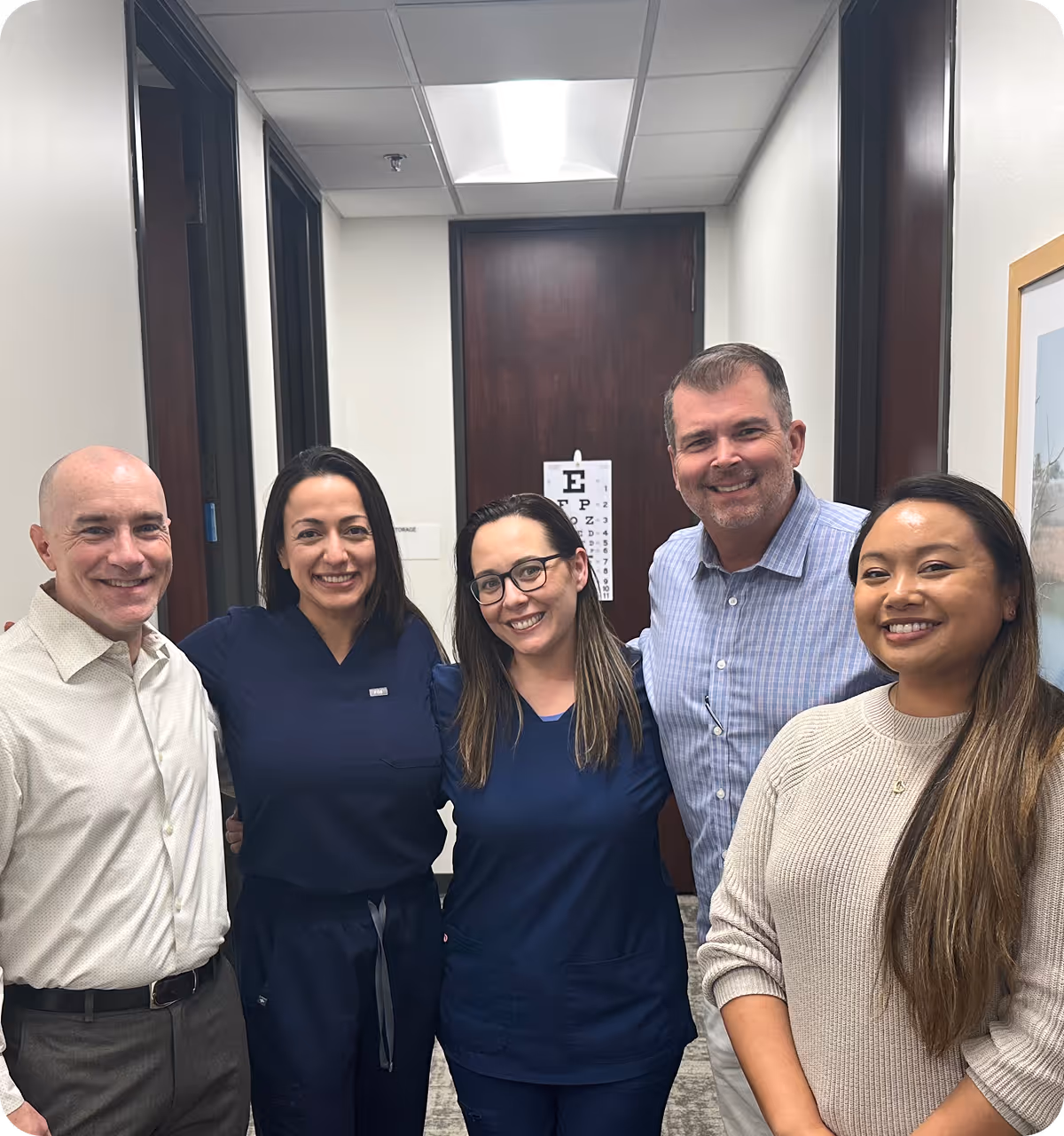 Five smiling adults standing in a hallway, two women in navy scrubs and others in casual or business attire.