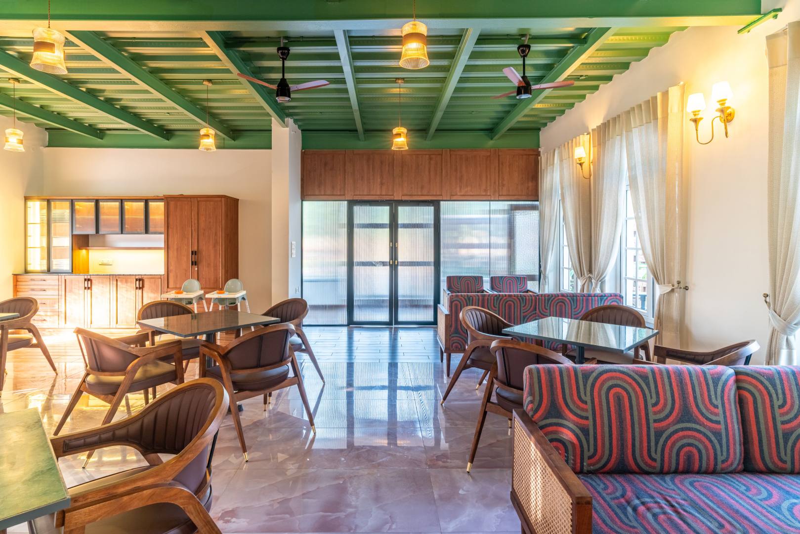 A warm and inviting modern dining hall at Sri Narayana Vanam farmhouse, featuring a unique green industrial-style slatted ceiling, polished marble floors, and elegant wooden furniture. The space is illuminated by warm hanging pendant lights and natural light from large windows with sheer drapes. Located at Survey number 129, Moinabad Mandal, Aziznagar, Hyderabad, Telangana 500075, this premium facility is perfect for group meals, corporate gatherings, and catering during farmhouse events.