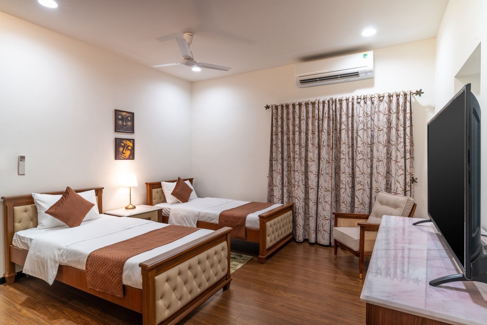 A comfortable guest bedroom at Sri Narayana Vanam farmhouse featuring twin beds with tufted cream headboards and brown accents. The room includes a flat-screen TV on a marble-top console, a classic armchair, and bamboo-patterned curtains. Located at Survey number 129, Moinabad Mandal, Aziznagar, Hyderabad, Telangana 500075, this accommodation is ideal for groups or families looking for a premium staycation.