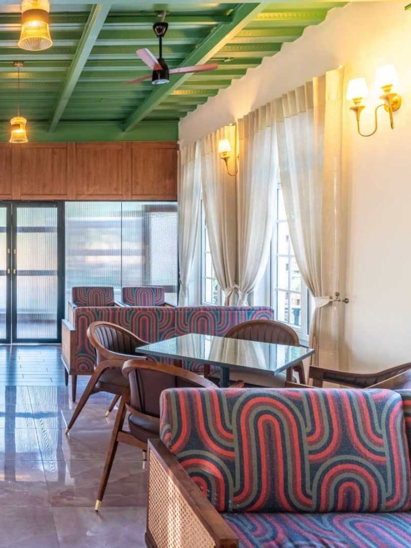 A warm and inviting modern dining hall at Sri Narayana Vanam farmhouse, featuring a unique green industrial-style slatted ceiling, polished marble floors, and elegant wooden furniture. The space is illuminated by warm hanging pendant lights and natural light from large windows with sheer drapes. Located at Survey number 129, Moinabad Mandal, Aziznagar, Hyderabad, Telangana 500075, this premium facility is perfect for group meals, corporate gatherings, and catering during farmhouse events.