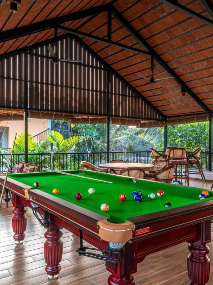 A spacious, high-ceilinged recreation lounge at Sri Narayana Vanam featuring a professional snooker table, polished wood-finish flooring, and a traditional terracotta-tiled roof. The area includes elegant cane-backed seating with floral cushions, perfect for social gatherings. This serene evening setting is located at Survey number 129, Moinabad Mandal, Aziznagar, Hyderabad, Telangana 500075, offering a peaceful atmosphere for staycation guests.