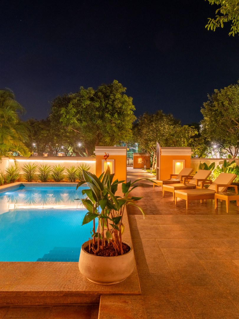 A stunning night view of the illuminated private swimming pool at Sri Narayana Vanam luxury farmhouse, featuring glowing underwater lights, premium poolside sun loungers, and warm ambient wall lighting. This tranquil vacation retreat is located at Survey number 129, Moinabad Mandal, Aziznagar, Hyderabad, Telangana 500075, offering a perfect setting for peaceful evening relaxation or intimate poolside celebrations.