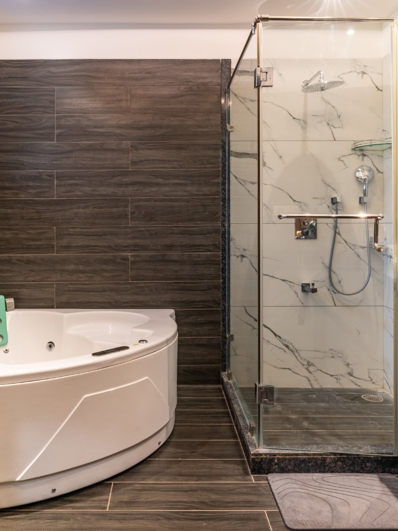 Modern luxury bathroom at Sri Narayana Vanam farmhouse featuring a large white corner bathtub and a separate glass-enclosed walk-in shower with premium marble tiling. Located at Survey number 129, Moinabad Mandal, Aziznagar, Hyderabad, Telangana 500075. A perfect high-end amenity for guests seeking a spa-like experience during their vacation rental stay.