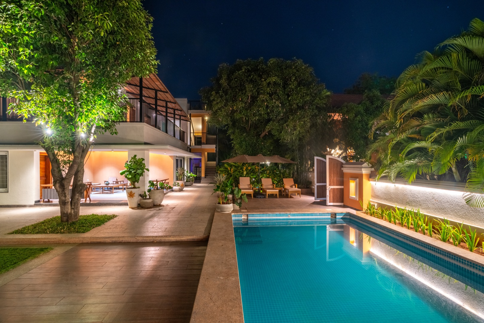 A night view of the illuminated private swimming pool at Sri Narayana Vanam luxury farmhouse, featuring a modern white villa with warm interior lighting. The poolside area includes premium wooden decking, comfortable lounge chairs under an umbrella, and lush tropical landscaping with lit-up greenery. This serene evening setting is located at Survey number 129, Moinabad Mandal, Aziznagar, Hyderabad, Telangana 500075, offering a peaceful atmosphere for staycation guests.