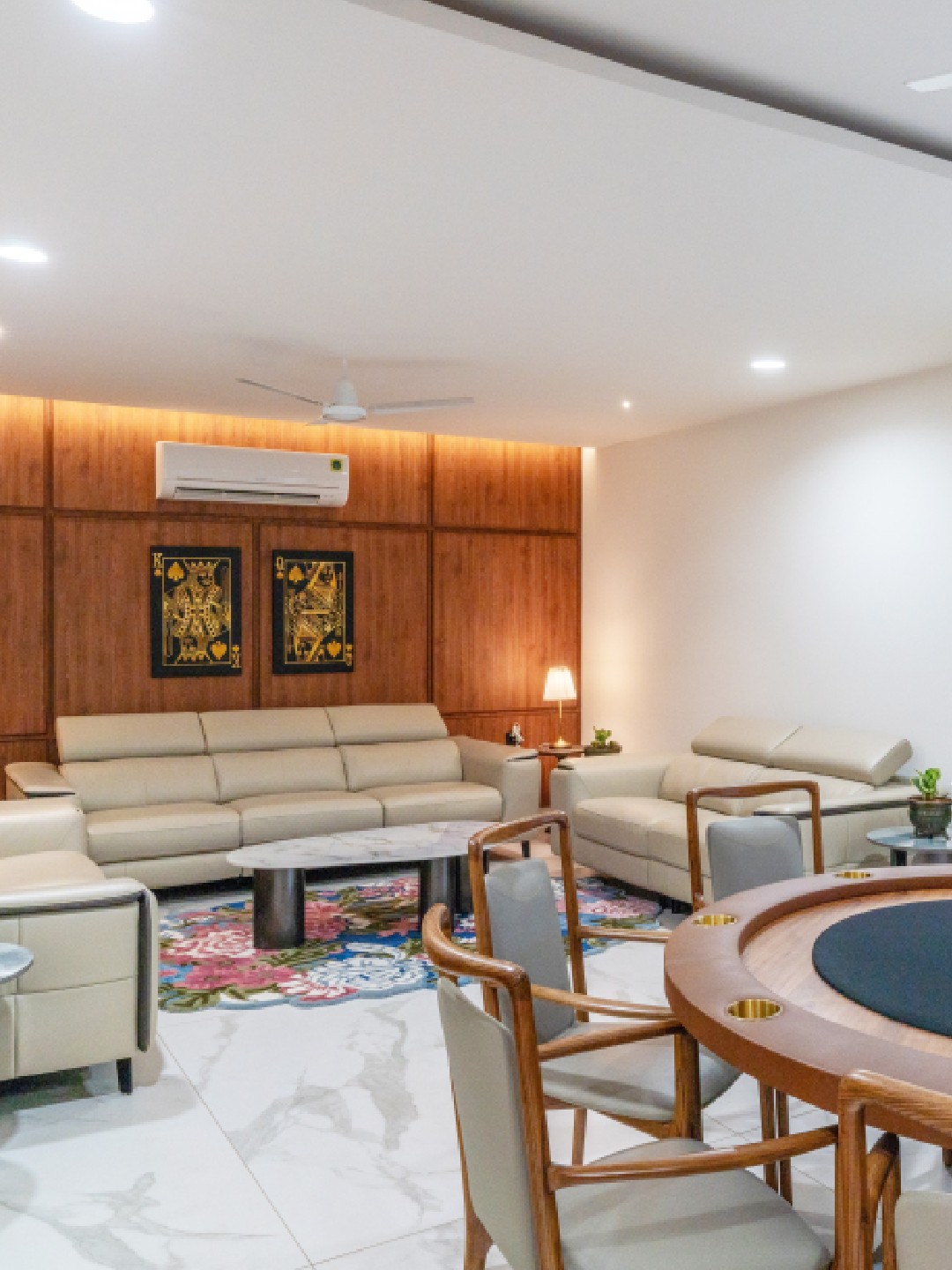 A sophisticated modern living room at Sri Narayana Vanam featuring a plush beige sectional sofa set against a warm, wood-paneled accent wall with integrated ambient lighting. The room includes a unique circular wooden card or gaming table with built-in cup holders, stylish marble-top side tables, and large glass doors with elegant neutral curtains. Located at Survey number 129, Moinabad Mandal, Aziznagar, Hyderabad, Telangana 500075, this luxury indoor space is perfectly designed for high-end social gatherings and private entertainment.