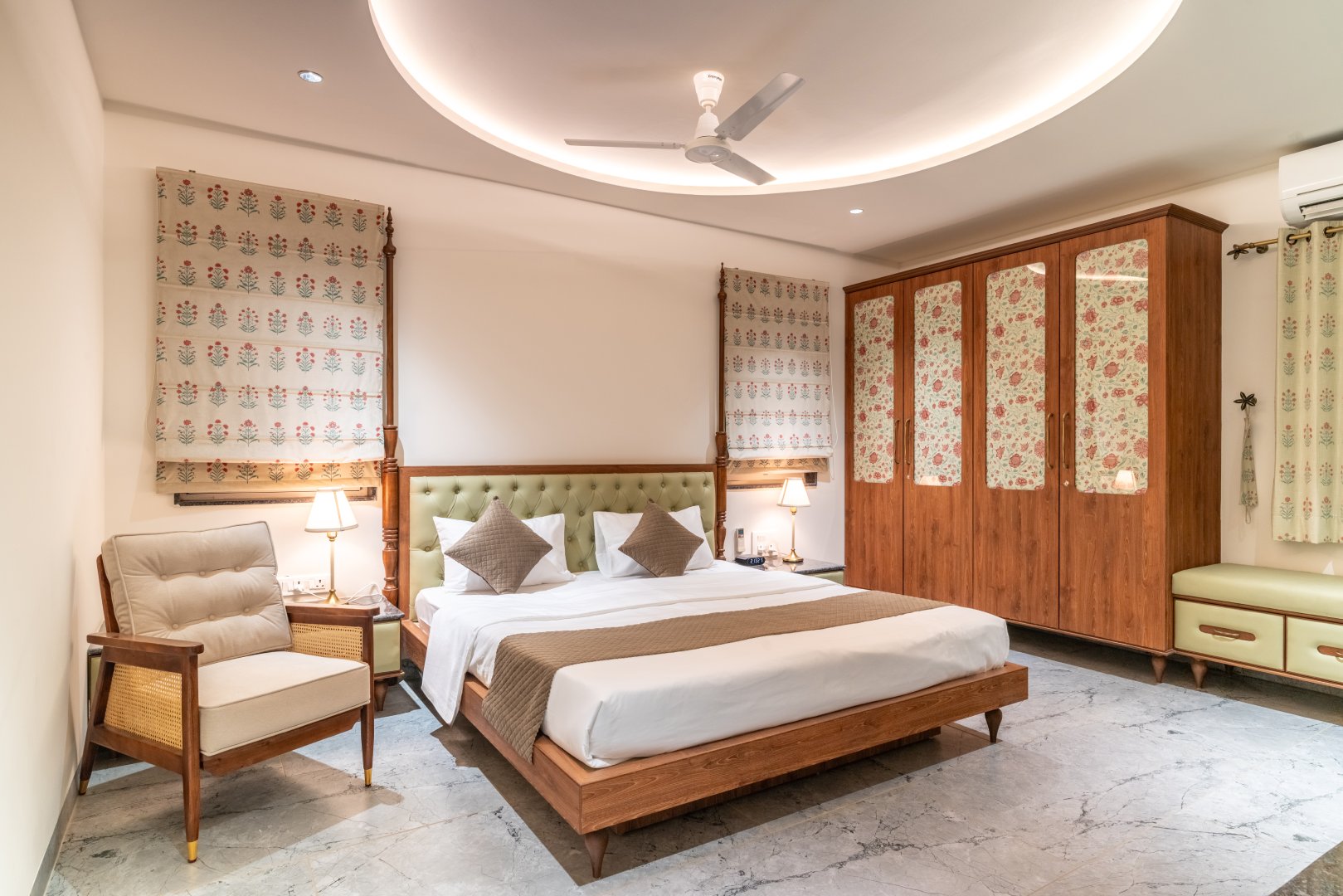 A luxury guest bedroom at Sri Narayana Vanam farmhouse featuring a king-size bed with a tufted sage-green headboard, elegant floral-patterned wardrobes, and a large circular recessed ceiling light. Located at Survey number 129, Moinabad Mandal, Aziznagar, Hyderabad, Telangana 500075. This premium accommodation offers a blend of classic and modern interior design for a comfortable vacation stay.