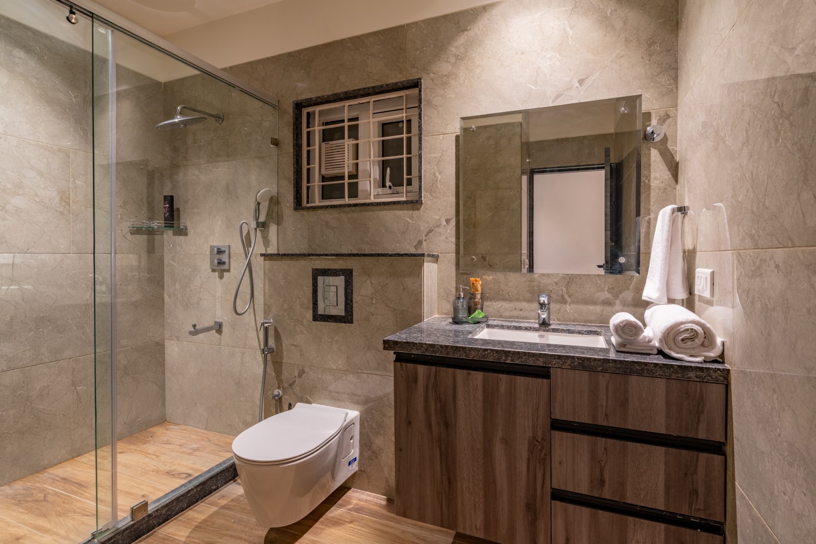 Spacious luxury bathroom at Sri Narayana Vanam farmhouse with a walk-in glass shower, modern soaking bathtub, granite countertop vanity, elegant wooden cabinets, and warm ambient lighting, part of a premium farm stay and vacation rental in Aziznagar, Moinabad, Hyderabad, located at Survey No. 129. Contact: 998-994-1555.