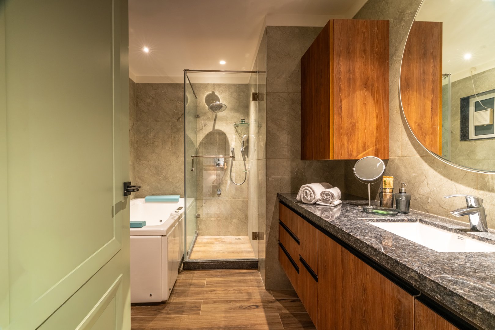 Luxury bathroom at Sri Narayana Vanam farmhouse featuring a walk-in glass shower, modern bathtub, premium granite vanity, wooden cabinetry, and warm ambient lighting, offering a high-end stay experience at a private farm stay in Aziznagar, Moinabad, Hyderabad, located at Survey No. 129. Contact: 998-994-1555.