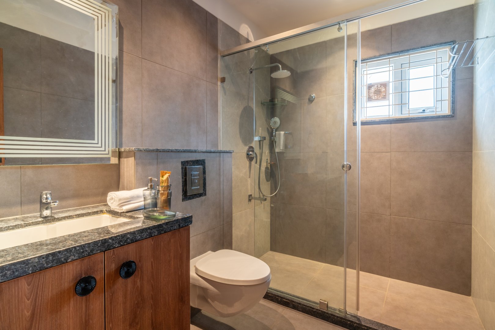 Luxury bathroom at Sri Narayana Vanam farmhouse featuring a walk-in glass shower, premium granite vanity, and modern grey tiling. This high-end amenity is part of our vacation rentals in Aziznagar, Moinabad, providing a refreshing experience for guests. Located at Survey number 129, Hyderabad. Phone: 998-994-1555.