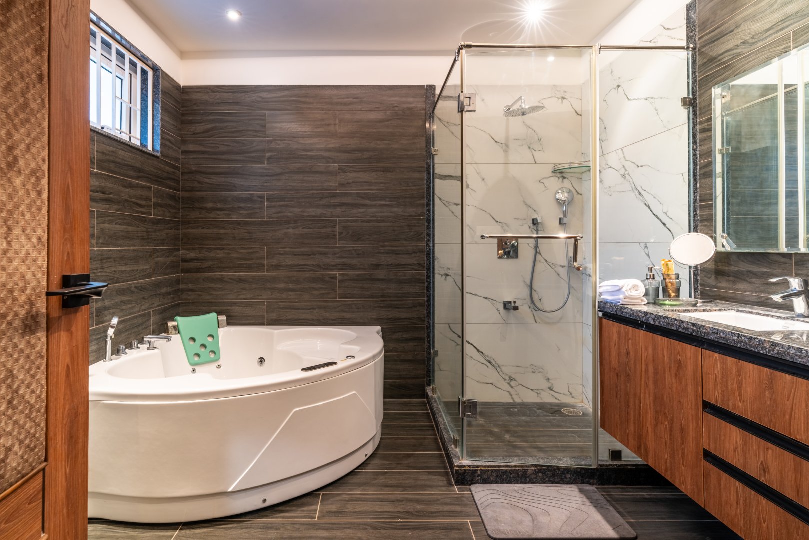 Luxury bathroom at Sri Narayana Vanam farmhouse featuring a spacious jacuzzi-style bathtub, walk-in glass shower with marble-finish tiles, granite vanity countertop, wooden cabinetry, and warm ambient lighting, offering a premium and relaxing experience at a private farm stay and vacation rental in Aziznagar, Moinabad, Hyderabad, located at Survey No. 129. Contact: 998-994-1555.