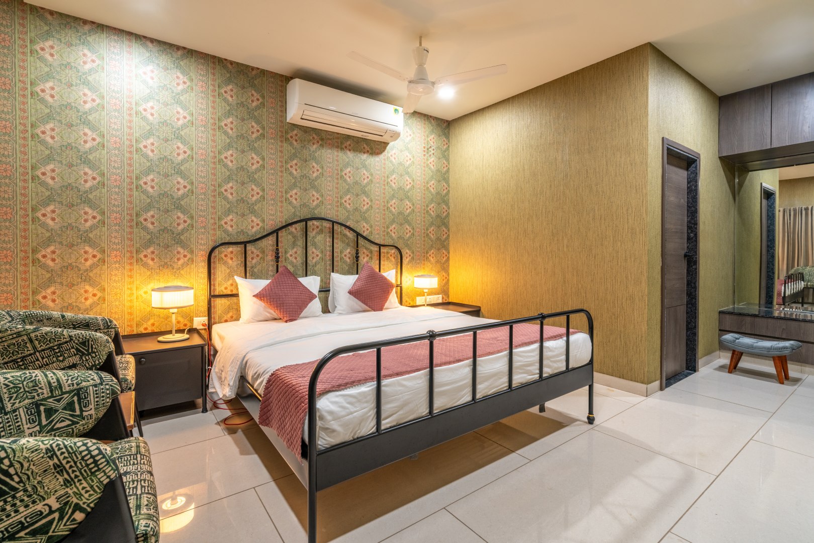 A contemporary guest bedroom at Sri Narayana Vanam featuring a metal-frame king-size bed with a patterned runner, set against an accent wall with intricate geometric wallpaper. The room includes warm bedside lighting, integrated air conditioning, and a vanity area with a cushioned stool. Located at Survey number 129, Moinabad Mandal, Aziznagar, Hyderabad, Telangana 500075, this suite offers a blend of modern comfort and elegant design for staycation guests.