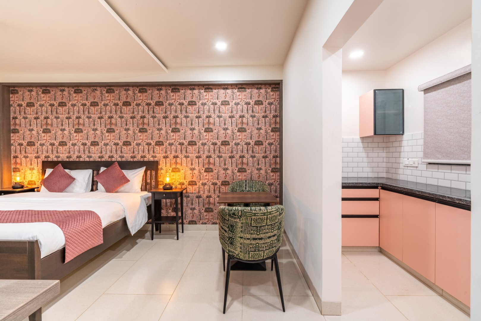 An elegant modern bedroom at Sri Narayana Vanam featuring a king-size bed with a dark wood frame, white linens, and pink accent pillows, set against a vibrant pink-patterned botanical wallpaper. The suite includes an integrated kitchenette with blush-pink cabinetry and white subway tile backsplash, a small dining table with green-patterned chairs, and warm bedside lamp lighting. Located at Survey number 129, Moinabad Mandal, Aziznagar, Hyderabad, Telangana 500075, this studio-style accommodation provides a luxurious and self-contained stay for guests.