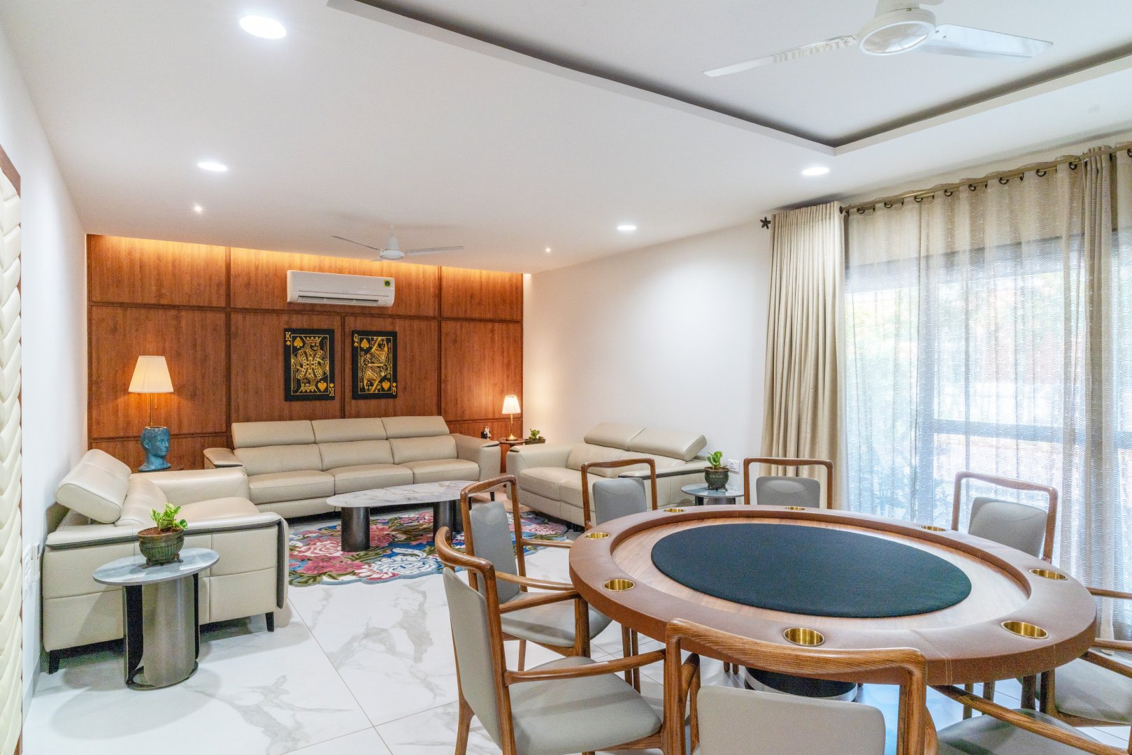 A sophisticated modern living room at Sri Narayana Vanam featuring a plush beige sectional sofa set against a warm, wood-paneled accent wall with integrated ambient lighting. The room includes a unique circular wooden card or gaming table with built-in cup holders, stylish marble-top side tables, and large glass doors with elegant neutral curtains. Located at Survey number 129, Moinabad Mandal, Aziznagar, Hyderabad, Telangana 500075, this luxury indoor space is perfectly designed for high-end social gatherings and private entertainment.