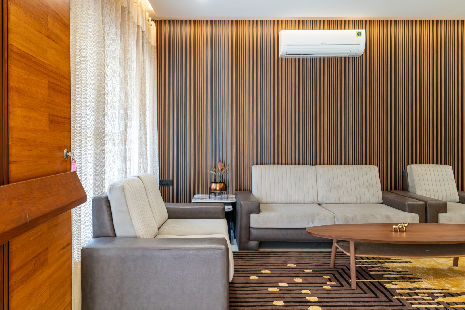 Modern luxury living room at Sri Narayana Vanam farmhouse featuring a plush cream and brown upholstered sofa set, contemporary slatted wood wall paneling, and an elegant marble-top side table. Located at Survey number 129, Moinabad Mandal, Aziznagar, Hyderabad, Telangana 500075. This stylish indoor lounge area offers a comfortable space for guests to relax during their stay.