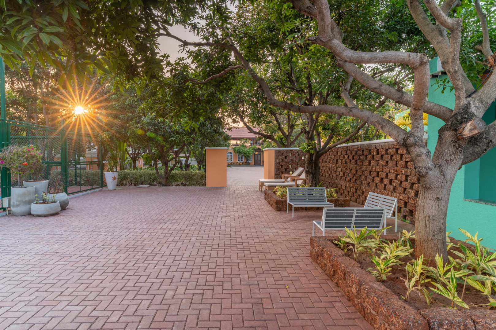 Scenic brick-paved outdoor courtyard at Sri Narayana Vanam farmhouse in Moinabad, Hyderabad, featuring a decorative red brick lattice wall, metal garden benches, and a sunburst flare through mature trees at Survey number 129, Aziznagar, Telangana 500075.