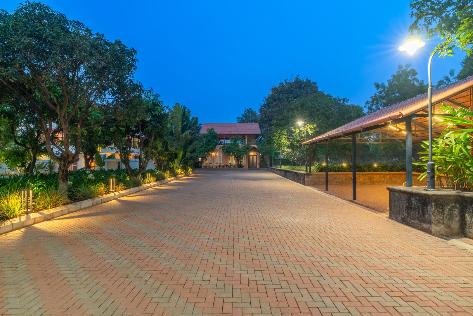Panoramic view of the expansive brick-paved driveway at Sri Narayana Vanam farmhouse in Moinabad, Hyderabad, leading to the main villa under a twilight sky with warm decorative street lamps and a covered parking bay at Survey number 129, Aziznagar, Telangana 500075.
