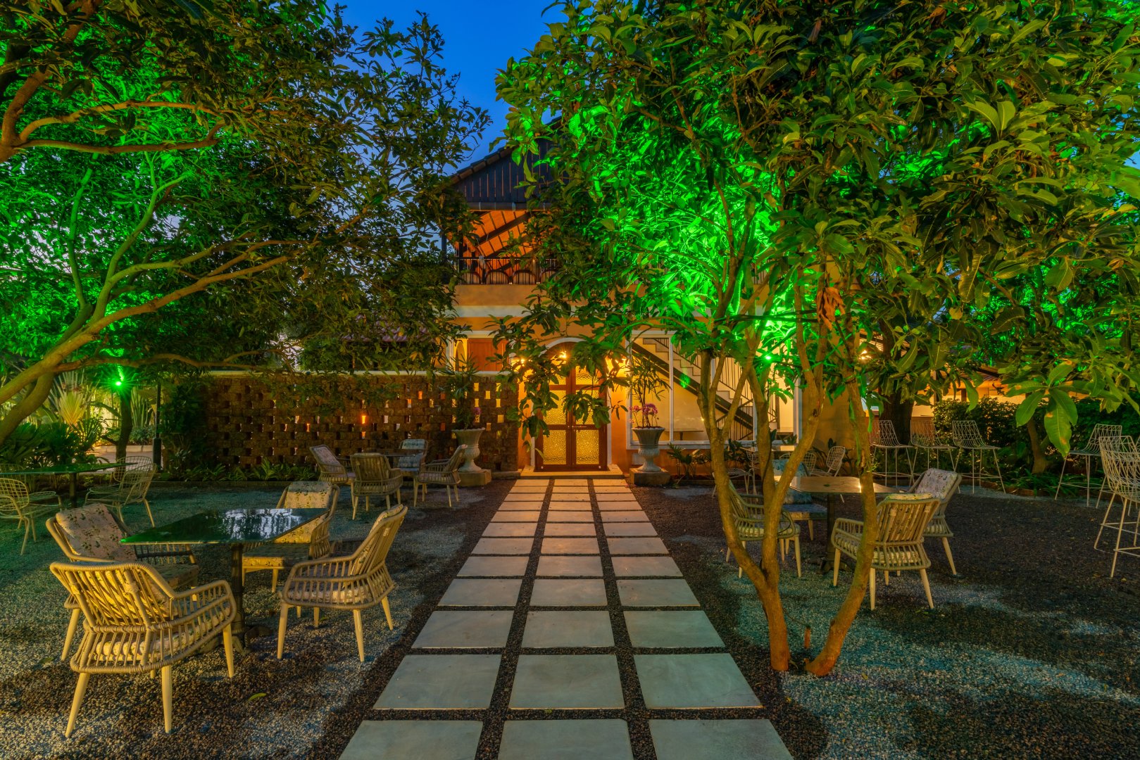 Elegant evening garden seating area at Sri Narayana Vanam farmhouse in Moinabad, Hyderabad, featuring a symmetrical stone tile walkway, premium wicker dining sets, and lush trees illuminated with atmospheric green lighting at Survey number 129, Aziznagar, Telangana 500075.