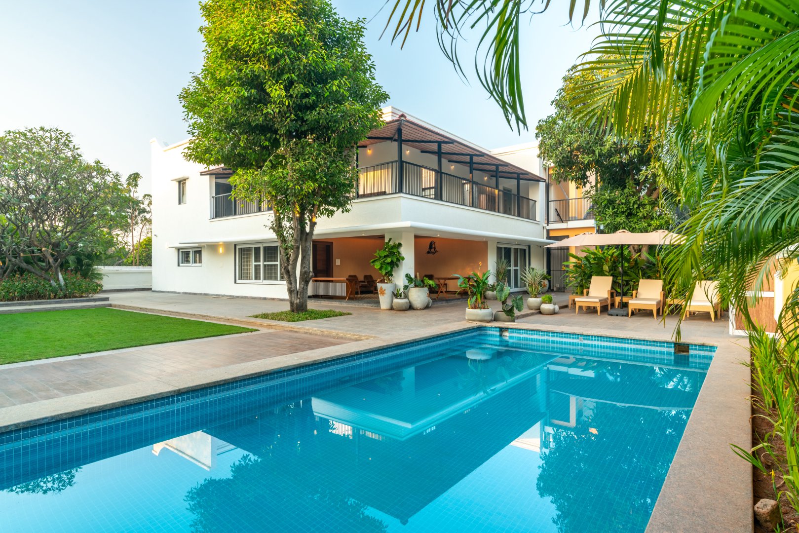 A crystal-clear blue swimming pool at Sri Narayana Vanam luxury farmhouse, reflecting the modern white villa architecture. The scene includes a poolside patio with potted plants, a sun lounger, and a spacious balcony. Located at Survey number 129, Moinabad Mandal, Aziznagar, Hyderabad, Telangana 500075.