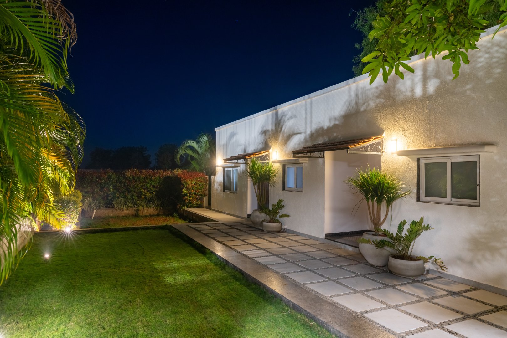 A beautifully lit outdoor walkway at Sri Narayana Vanam farmhouse in Moinabad, Hyderabad, featuring checkered stone paving, white exterior walls with warm wall-mounted lamps, and a manicured green lawn at Survey number 129, Aziznagar, Telangana 500075.