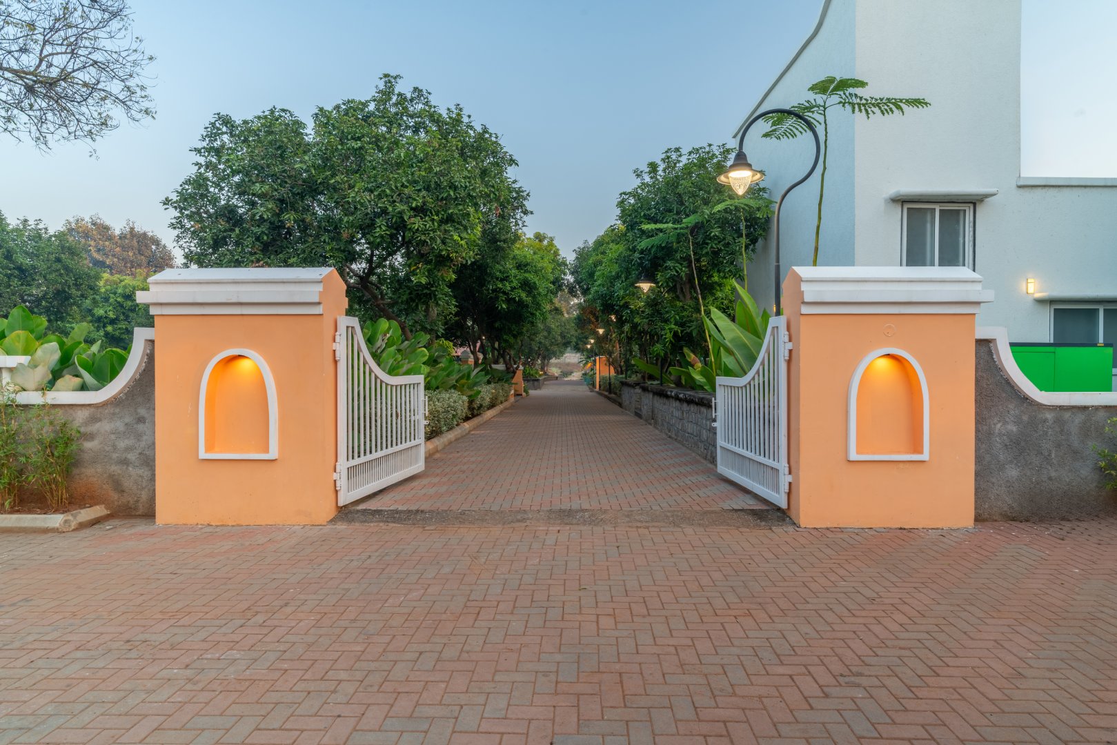 Modern semi-outdoor pavilion at Sri Narayana Vanam farmhouse in Moinabad, Hyderabad, featuring comfortable white armchairs, decorative hanging light fixtures, and a green accent wall overlooking the lush gardens at Survey number 129, Aziznagar, Telangana 500075.