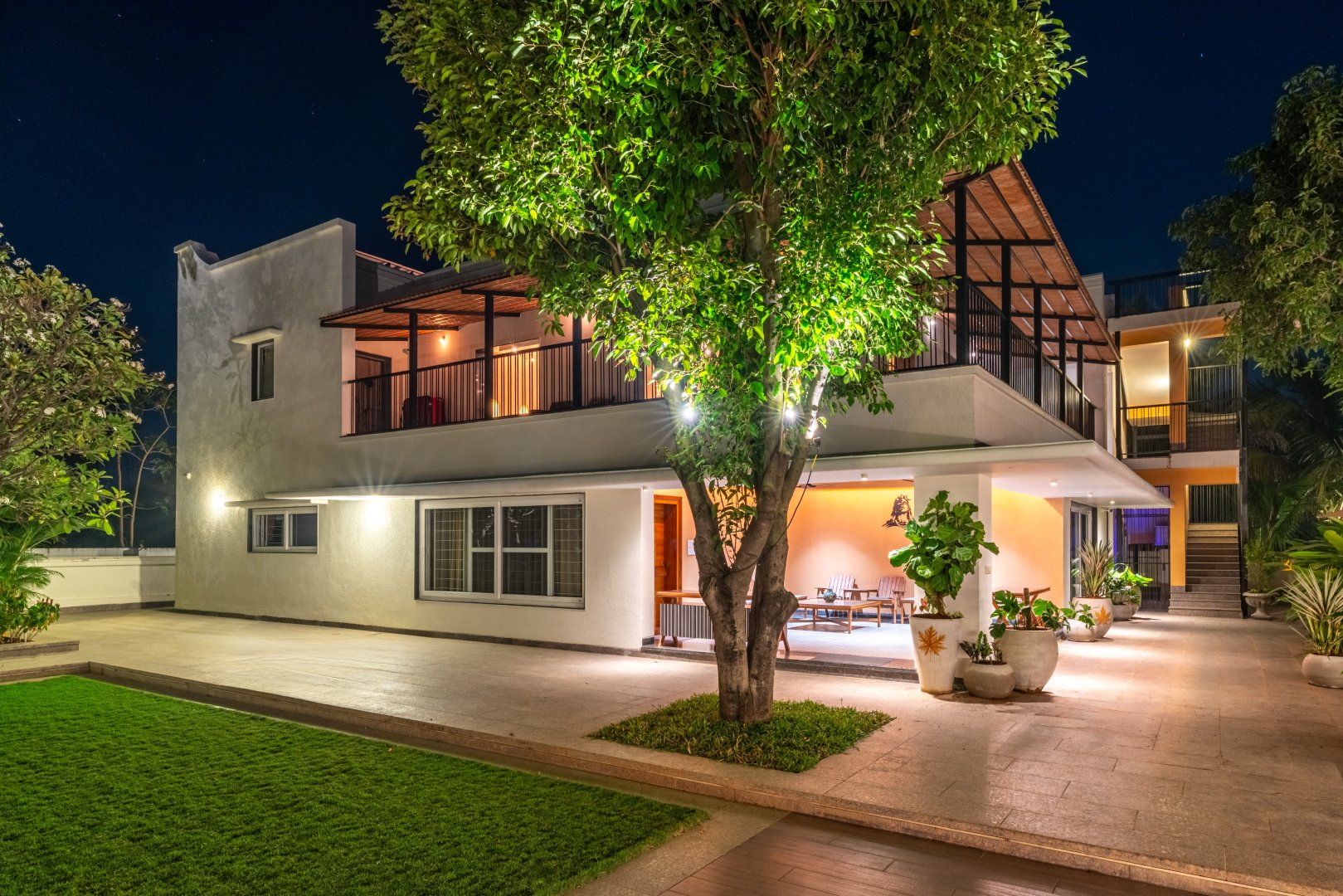 Modern semi-outdoor pavilion at Sri Narayana Vanam farmhouse in Moinabad, Hyderabad, featuring comfortable white armchairs, decorative hanging light fixtures, and a green accent wall overlooking the lush gardens at Survey number 129, Aziznagar, Telangana 500075.