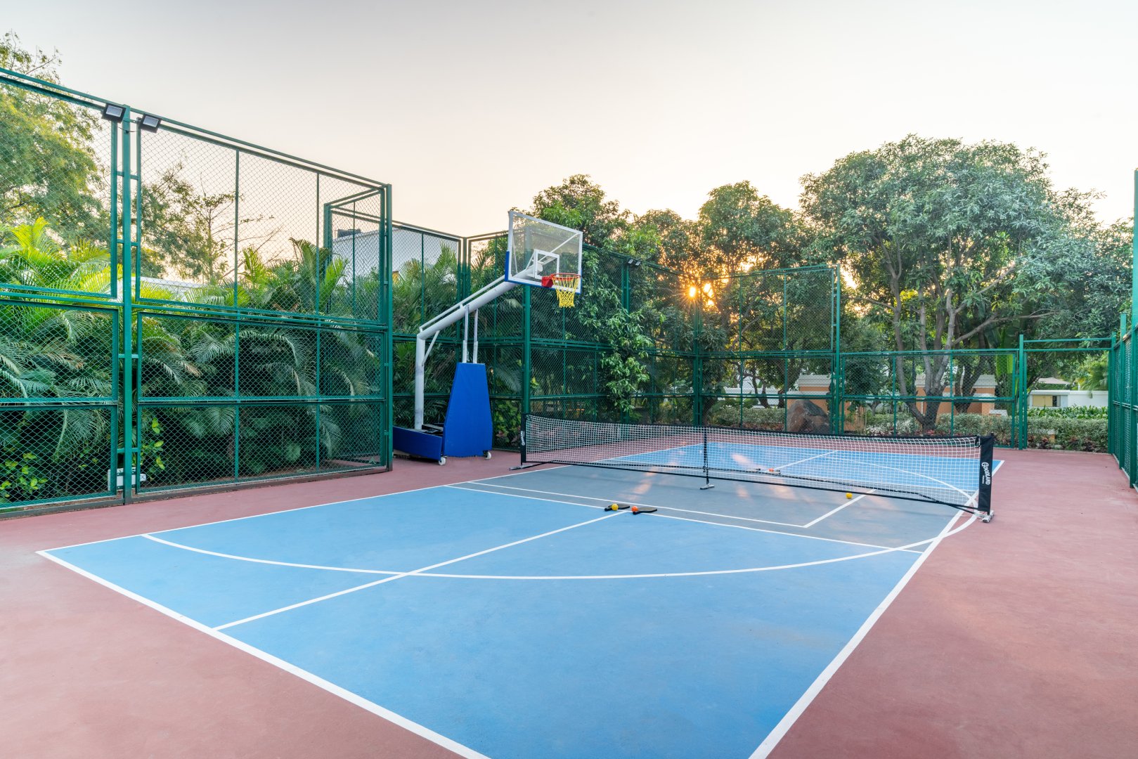 Professional outdoor multi-purpose sports court at Sri Narayana Vanam farmhouse in Moinabad, Hyderabad, featuring a basketball hoop and tennis net with high safety fencing and lush perimeter greenery at Survey number 129, Aziznagar, Telangana 500075.