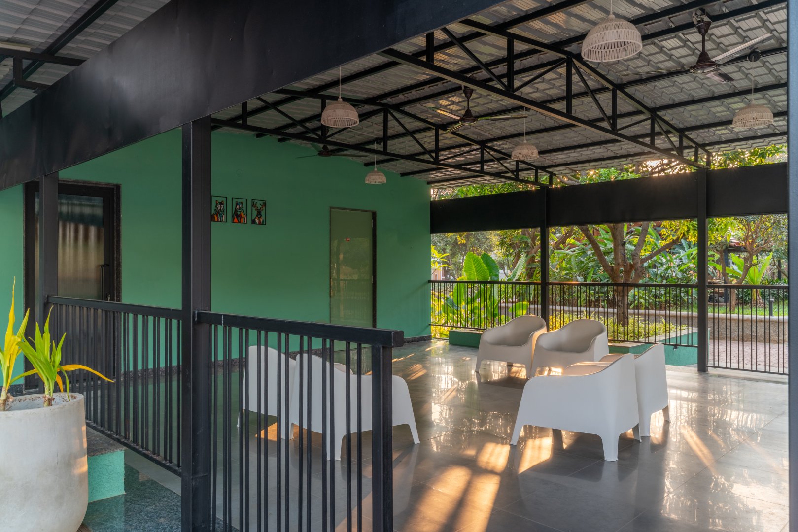 Modern semi-outdoor pavilion at Sri Narayana Vanam farmhouse in Moinabad, Hyderabad, featuring comfortable white armchairs, decorative hanging light fixtures, and a green accent wall overlooking the lush gardens at Survey number 129, Aziznagar, Telangana 500075.