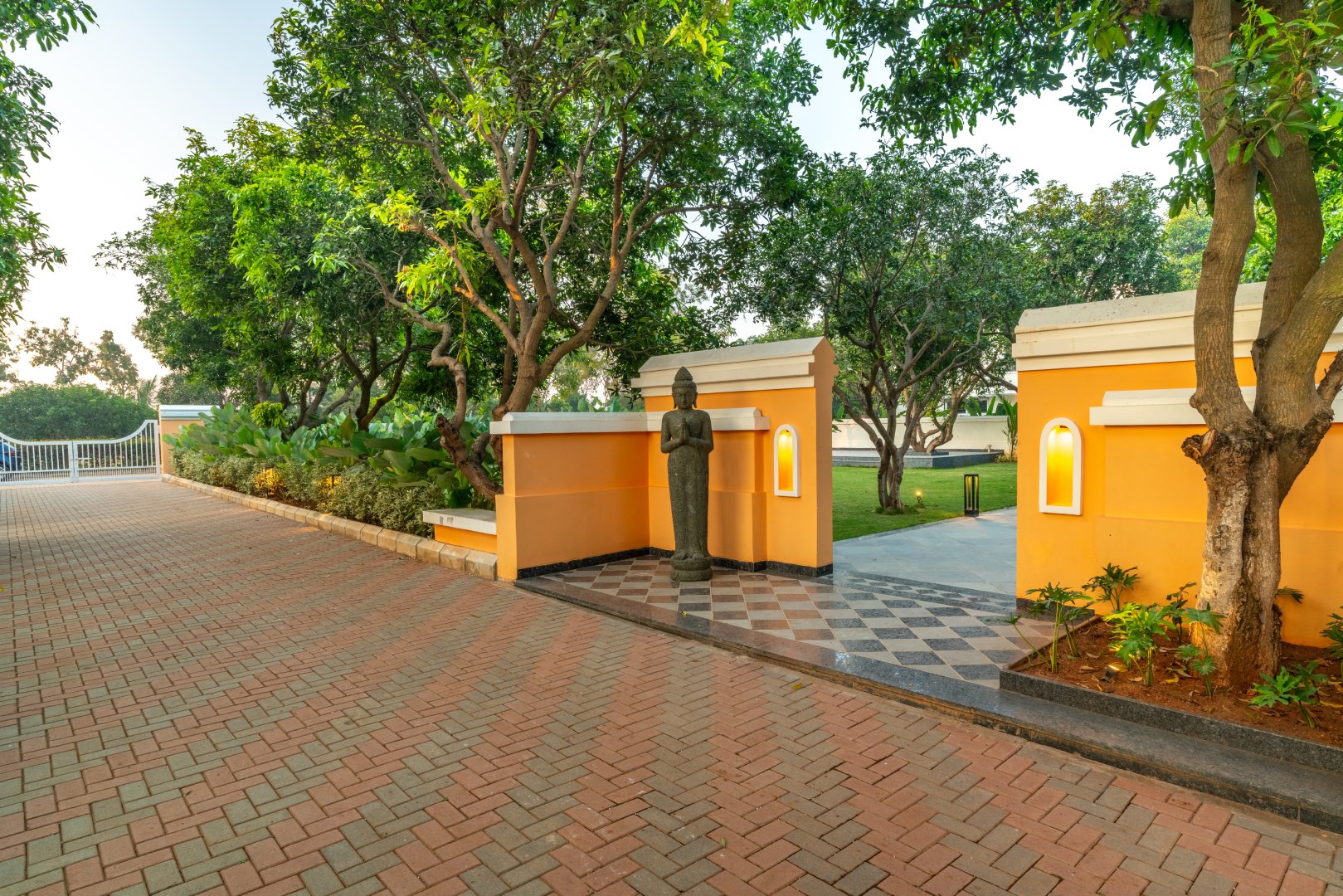 Daytime view of the grand entrance at Sri Narayana Vanam farmhouse in Moinabad, Hyderabad, featuring a traditional stone statue, yellow compound walls with inset lighting, and a paved brick driveway surrounded by lush greenery at Survey number 129, Aziznagar, Telangana 500075.