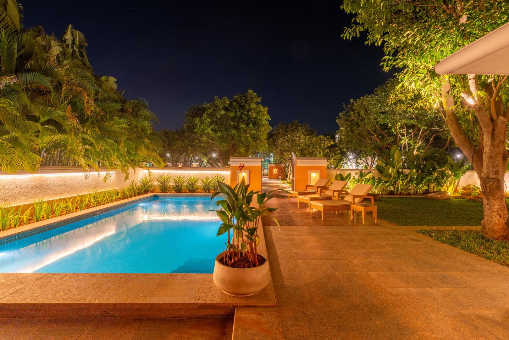 A serene nighttime view of the private swimming pool at Sri Narayana Vanam luxury farmhouse in Aziznagar, Hyderabad. The pool is beautifully illuminated with glowing blue water, surrounded by a paved deck, lush green lawns, and warm landscape lighting on the trees and perimeter walls. Located at Survey number 129, Moinabad Mandal, Aziznagar, Hyderabad, Telangana 500075, this peaceful outdoor setting is perfect for evening relaxation during a premium staycation.