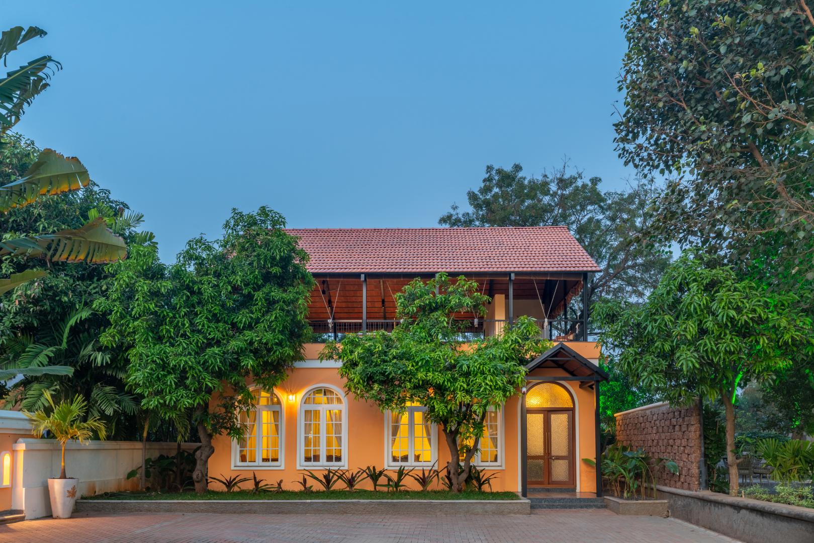 Exterior view of a charming holiday cottage guest room at a private luxury farm stay. This boutique retreat features warm lighting, classic arched windows, and lush greenery at Survey number 129, Moinabad Mandal, Aziznagar, Hyderabad, Telangana 500075. An ideal vacation rental for couples seeking peace and privacy.