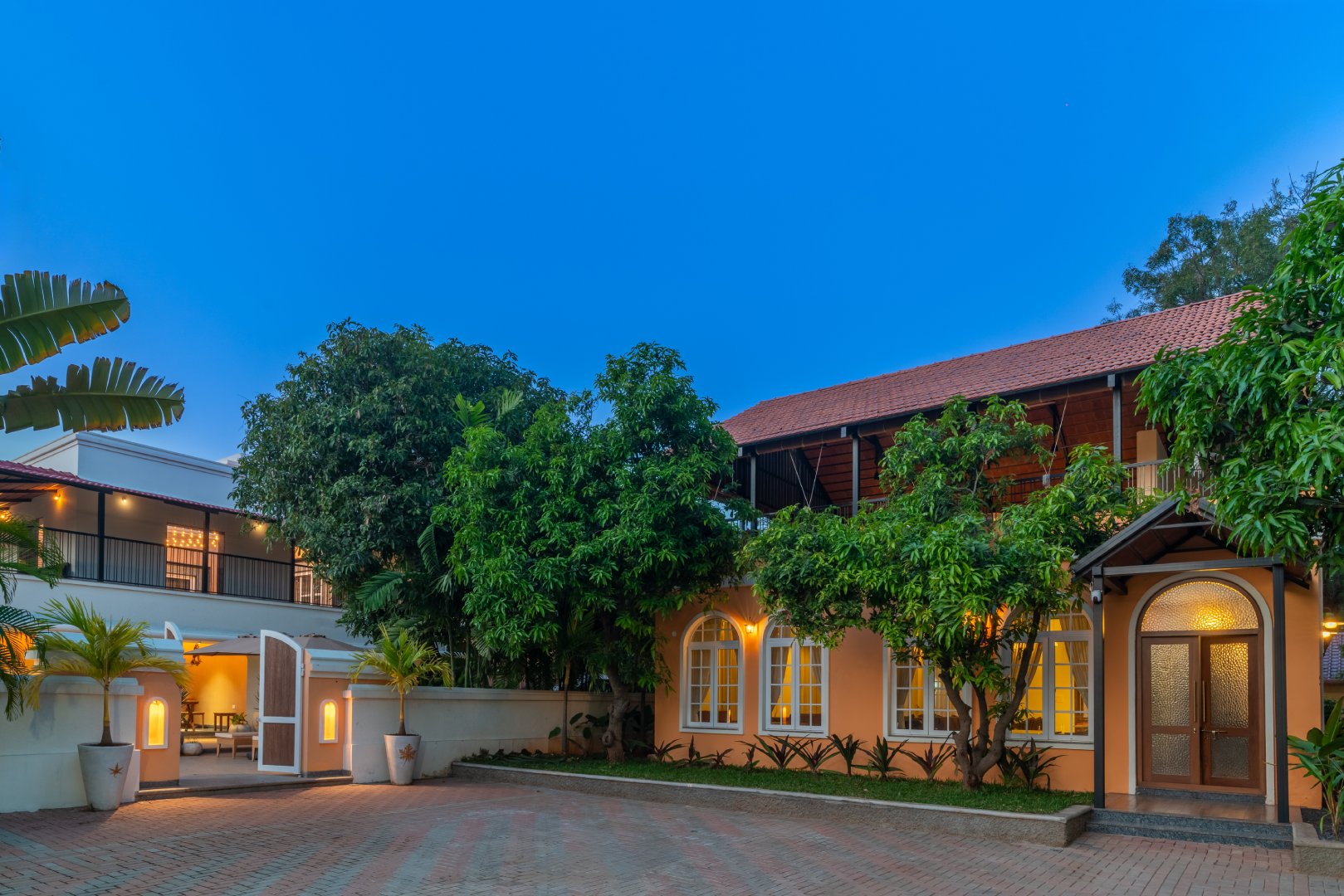Exterior evening view of the main luxury villa at Sri Narayana Vanam farmhouse in Moinabad, Hyderabad, showing the arched windows, tiled roof, and beautifully landscaped paved entrance at Survey number 129, Aziznagar, Telangana 500075.