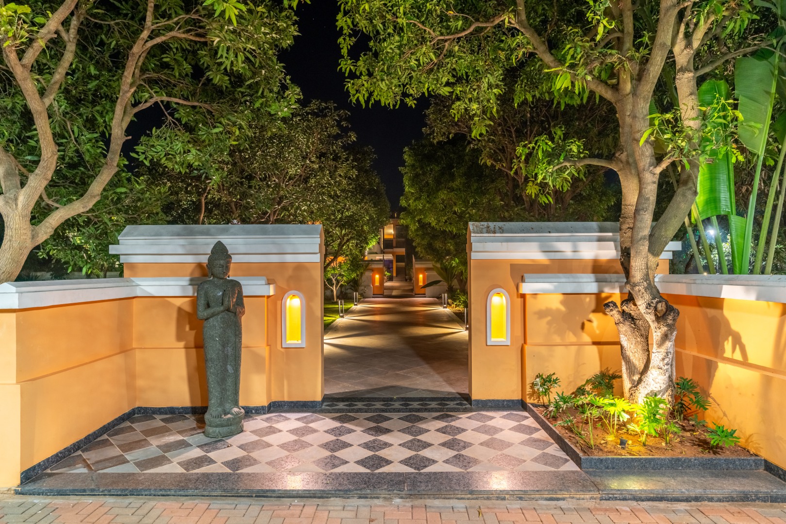 The elegant entrance of Sri Narayana Vanam farmhouse in Moinabad, Hyderabad, featuring a traditional stone statue in a welcoming pose, lit-up yellow walls, and a checkered stone pathway leading into the lush estate at Survey number 129, Aziznagar, Telangana 500075.