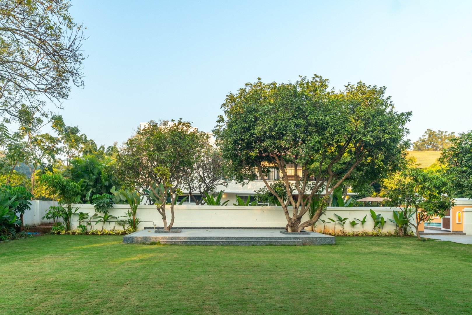 Expansive, manicured green lawn at Sri Narayana Vanam farmhouse in Moinabad, Hyderabad, featuring a raised stone platform under mature trees, perfect for outdoor events and garden parties at Survey number 129, Aziznagar, Telangana 500075.