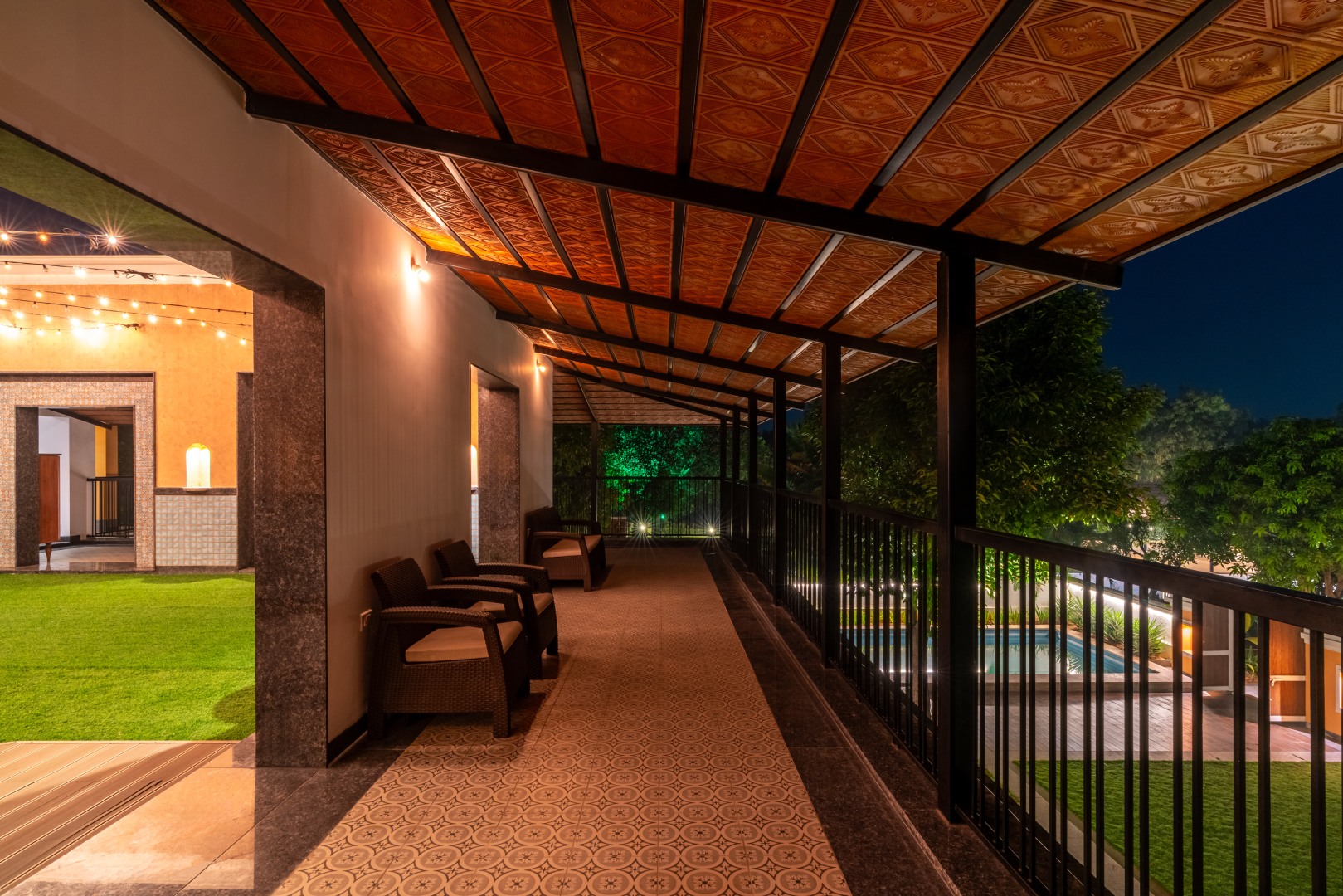 Illuminated outdoor corridor at Sri Narayana Vanam farmhouse in Moinabad, Hyderabad, featuring a rustic textured ceiling, comfortable wicker seating, and a view of the swimming pool and garden at night at Survey number 129, Aziznagar, Telangana 500075.