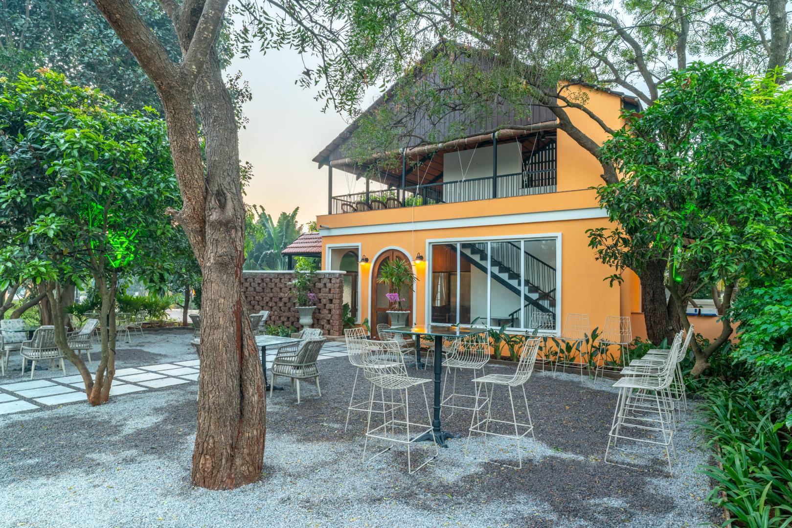 Exterior view of the two-story villa at Sri Narayana Vanam farmhouse in Moinabad, Hyderabad, featuring an orange facade, a spacious first-floor balcony with a sloping roof, and a shaded outdoor gravel seating area surrounded by lush trees at Survey number 129, Aziznagar, Telangana 500075.
