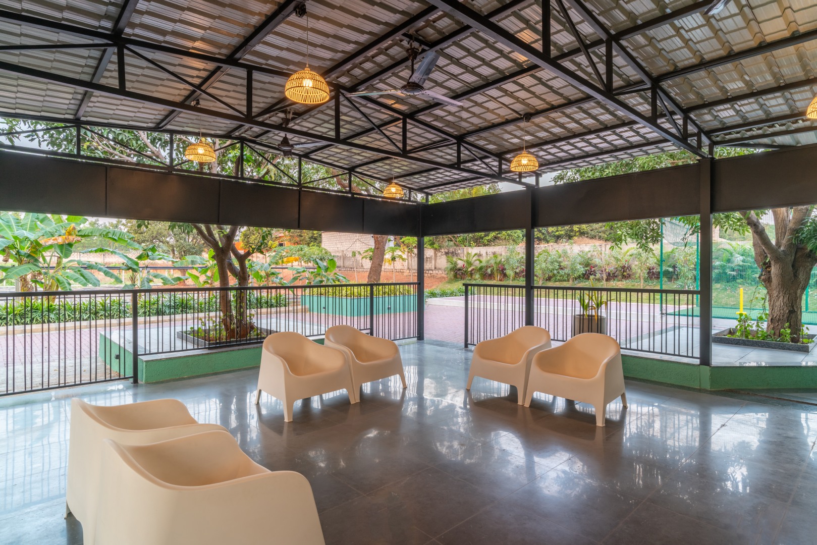 Modern open-air lounge at Sri Narayana Vanam farmhouse in Moinabad, Hyderabad, featuring comfortable white bucket chairs under a high-clearance metal roof with decorative hanging lights and views of the lush garden at Survey number 129, Aziznagar, Telangana 500075.