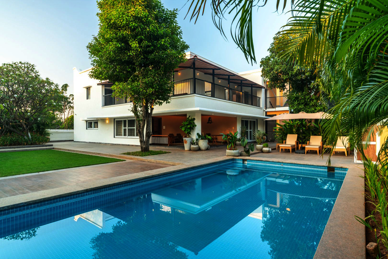 A vibrant day scene at Sri Narayana Vanam farmhouse showing families and children enjoying a large private swimming pool with floats, while others relax on the villa balconies and poolside deck. Located at Survey number 129, Moinabad Mandal, Aziznagar, Hyderabad, Telangana 500075, this luxury vacation rental offers the perfect setting for multi-generational family gatherings and lively poolside staycations.