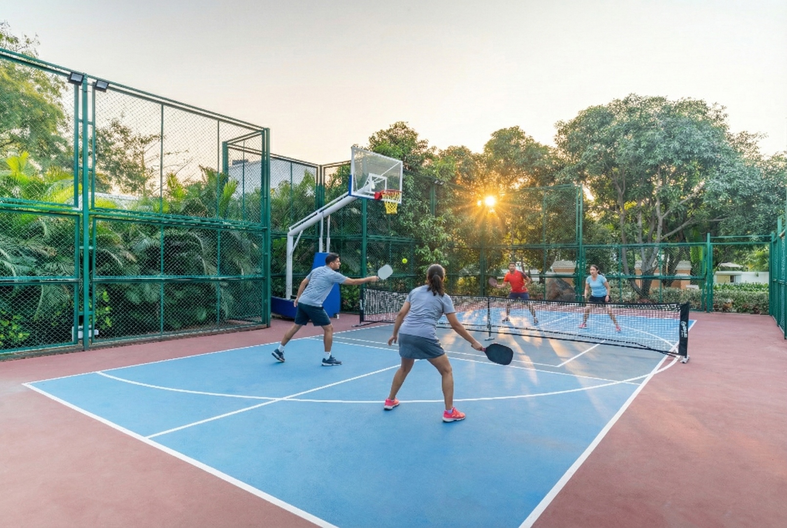 Professional outdoor pickleball and basketball court at Sri Narayana Vanam farmhouse, featuring guests playing a match during sunset on a high-quality blue and red sports surface. Located at Survey number 129, Moinabad Mandal, Aziznagar, Hyderabad, Telangana 500075. A premier amenity for sports enthusiasts, active vacations, and corporate team-building events in a lush nature setting.