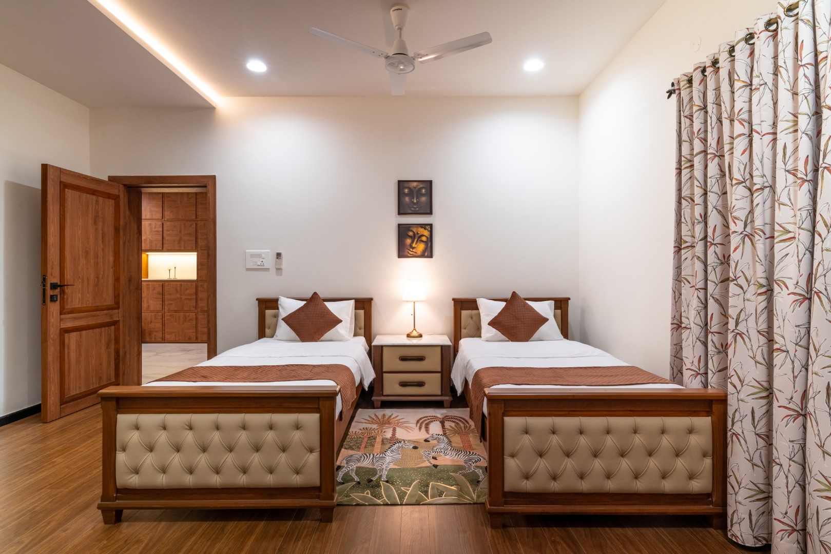 A luxury guest bedroom at Sri Narayana Vanam farmhouse featuring twin beds with tufted cream footboards, a vibrant animal-themed area rug, and an open doorway leading to a private dressing area. Located at Survey number 129, Moinabad Mandal, Aziznagar, Hyd