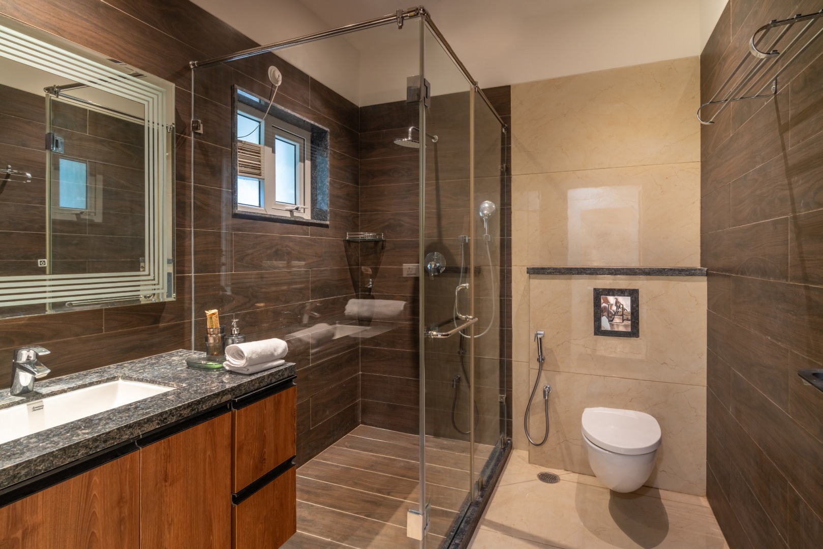 A luxury ensuite bathroom at Sri Narayana Vanam farmhouse featuring dark wood-textured wall tiles, a granite-top vanity with a wooden cabinet, and a large backlit mirror. The space includes a glass-enclosed walk-in shower and premium white sanitary ware. L