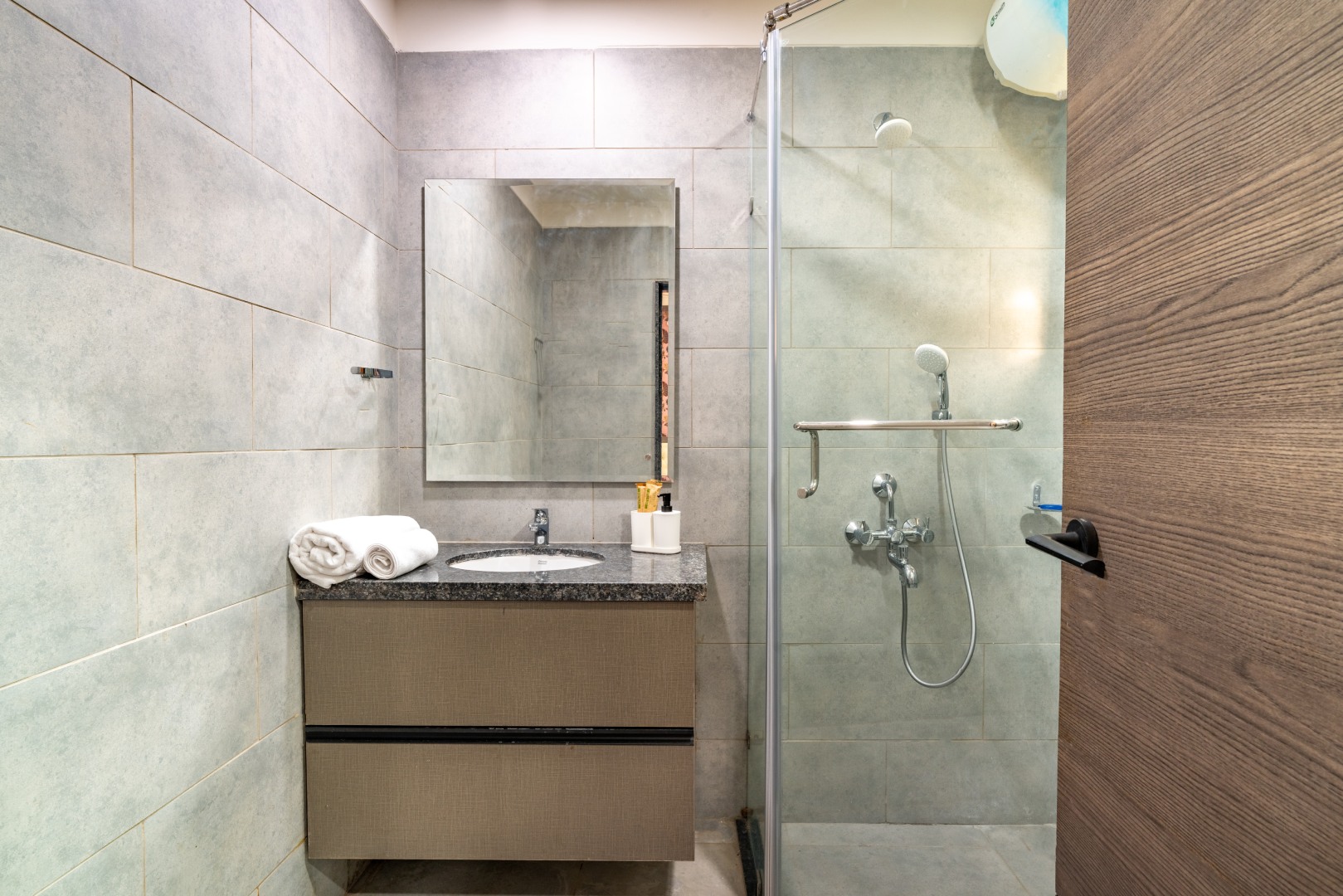 A sleek, modern ensuite bathroom at Sri Narayana Vanam farmhouse featuring grey stone-textured wall tiles, a floating wooden vanity with a black granite top, and a glass-enclosed walk-in shower. Located at Survey number 129, Moinabad Mandal, Aziznagar, Hyd
